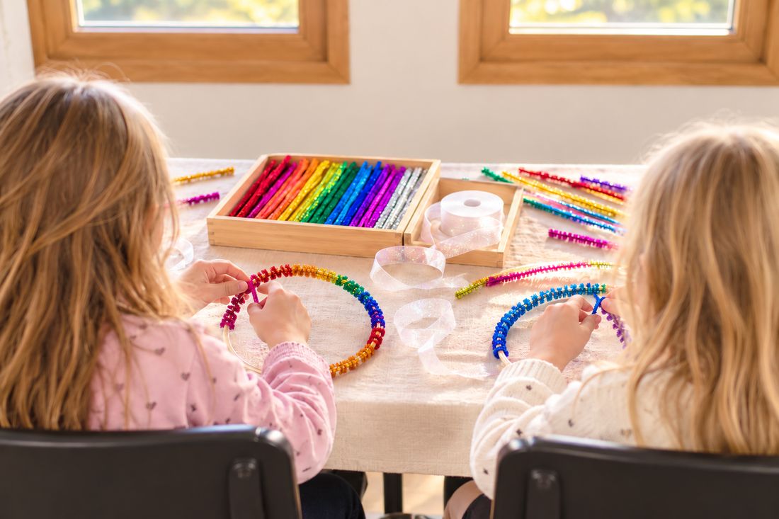 Zwei Mädchen basteln am Tisch Regenbogen-Ringe aus bunten Pfeifenputzern